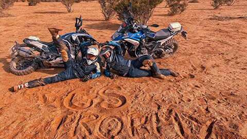 Two riders on the International women’s team lying in the sand in front of their motorcycles, with “GS Trophy” written in the sand.